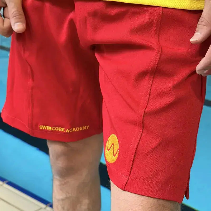 Yellow Swimcore Academy embroidery on the leg of the red lifeguard shorts