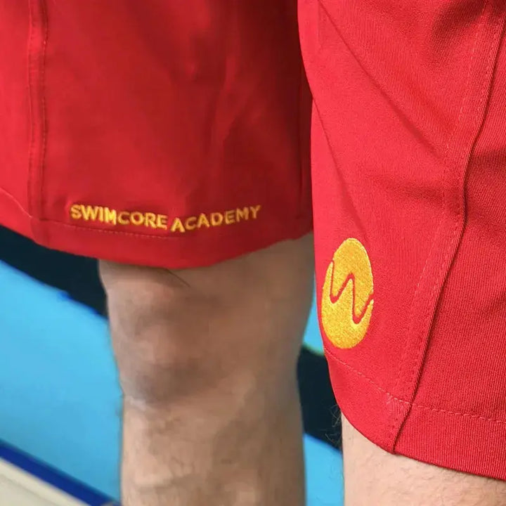 Embroidered Swimcore Academy text and wave logo on the red lifeguard shorts